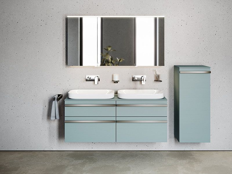 Waschtischanlage mit Waschtischunterschrank und daneben ein Hochschrank, beides in Eisblau. Auf dem Unterschrank zwei Waschbecken mit jeweils einem Unterputz Wasserhahn. Darüber ein beleuchteter Spiegelschrank. Die Möbel sind von FOR aus der Serie MyStyle.